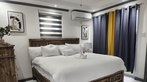 A neatly made bed with white linens and pillows, a wooden headboard, a bedside lamp, and a window with striped blinds.