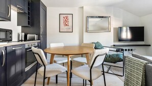 Eine moderne Küche mit dunklen Schränken, einer Mikrowelle und einem Herd. In der Mitte steht ein runder Holztisch mit Stühlen. An der Wand über einer Couch ist ein Flachbildfernseher angebracht, auf der Couch liegt ein dekoratives Gewebe.