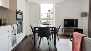 Una cocina moderna con electrodomésticos integrados, un comedor con mesa y sillas, un televisor de pantalla plana y vistas a un paisaje urbano a través de un ventanal grande.