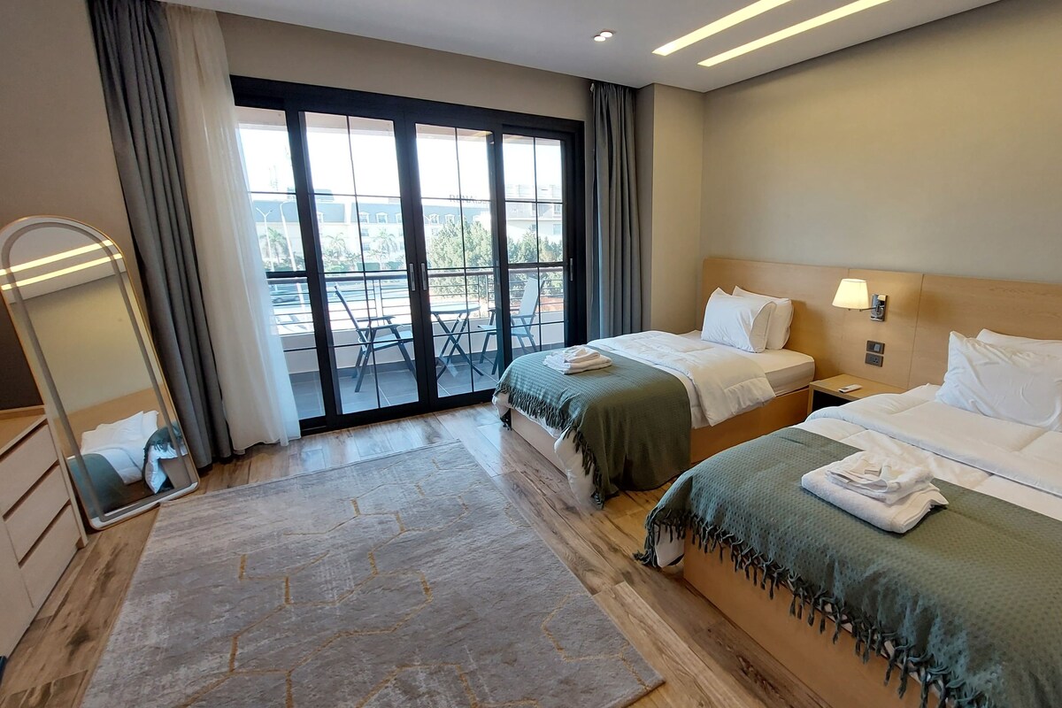 A hotel room with two beds, a large window with a view, a mirror, and a wooden floor.