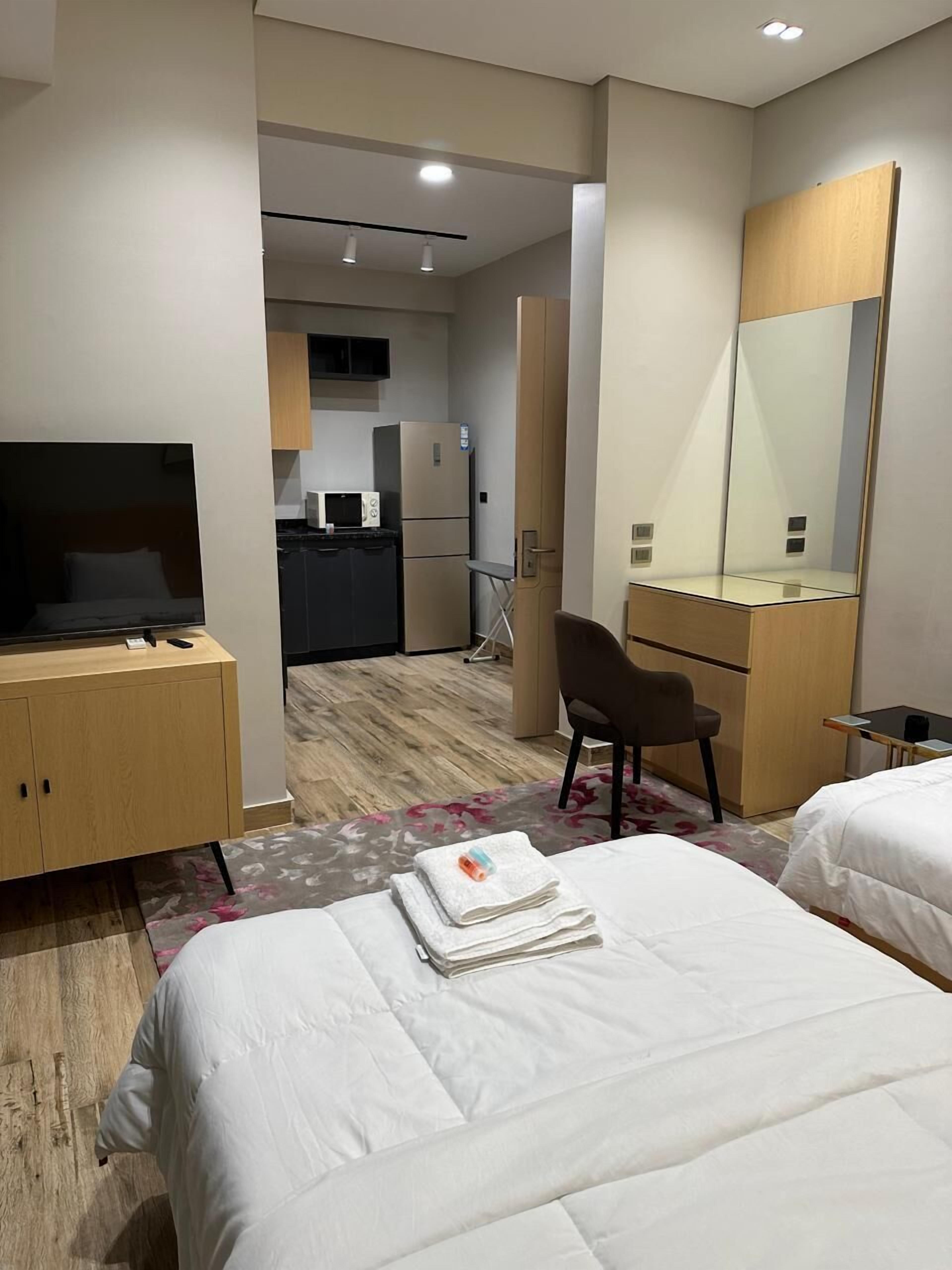 Habitación de hotel con dos camas, un escritorio con televisor, una silla, una pequeña cocina y un espejo grande.