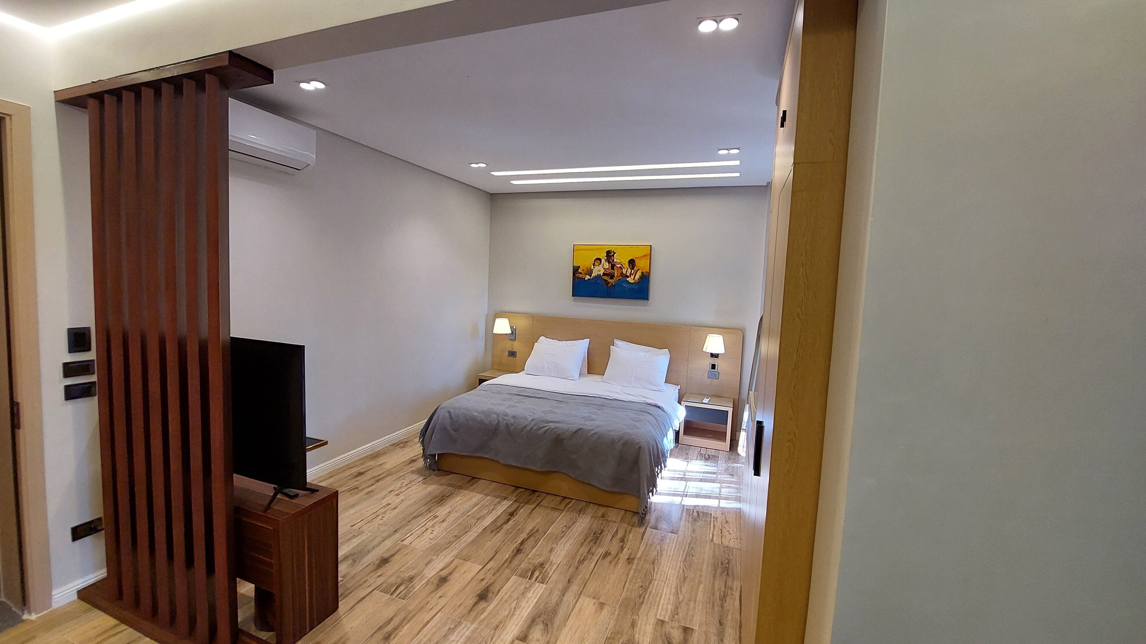 A modern hotel room with a wooden floor, a bed with white linens, a flat-screen TV, and a painting on the wall.