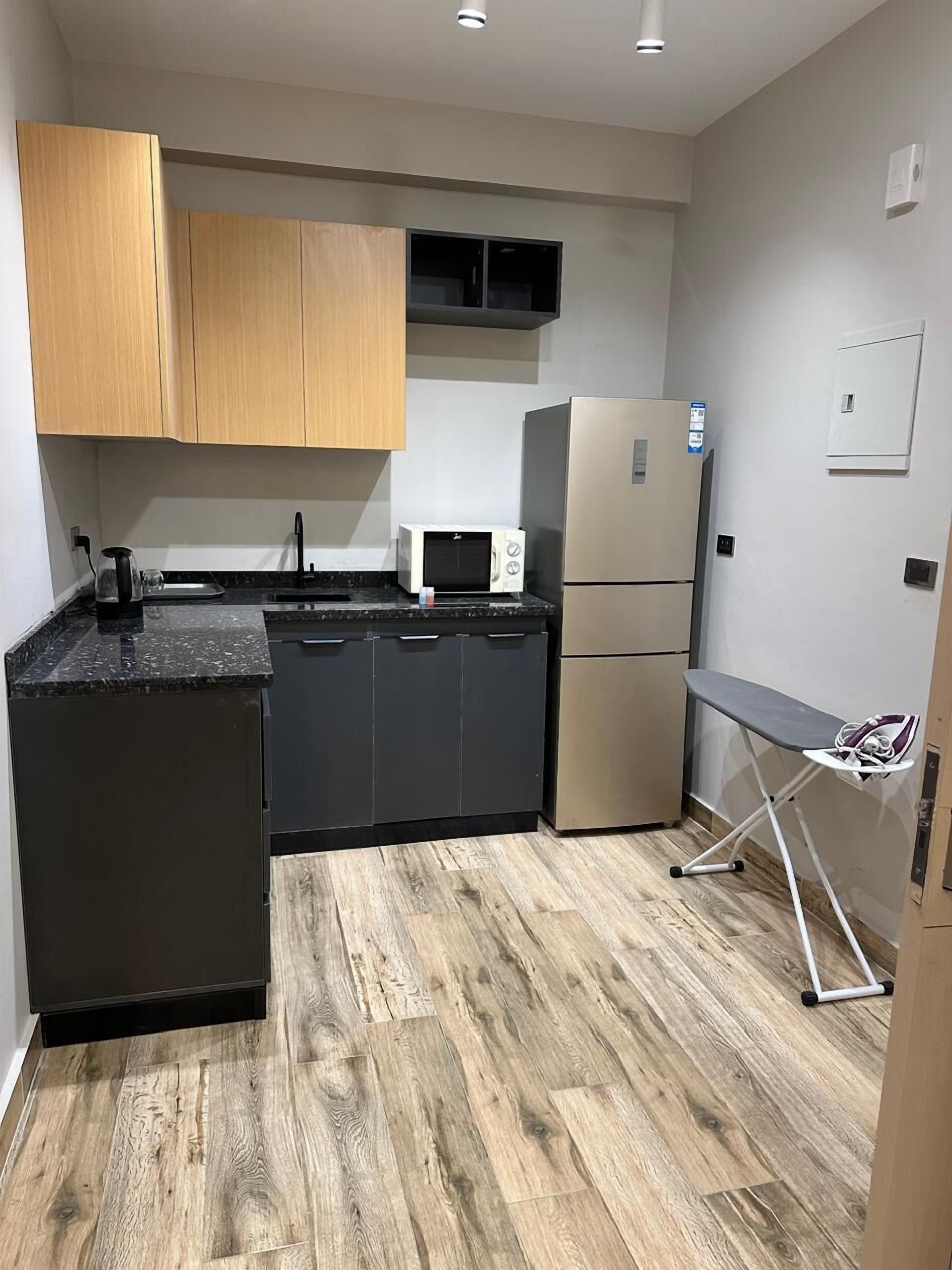 A compact kitchen with wooden cabinets, a black countertop, a microwave, a refrigerator, and a small ironing board.