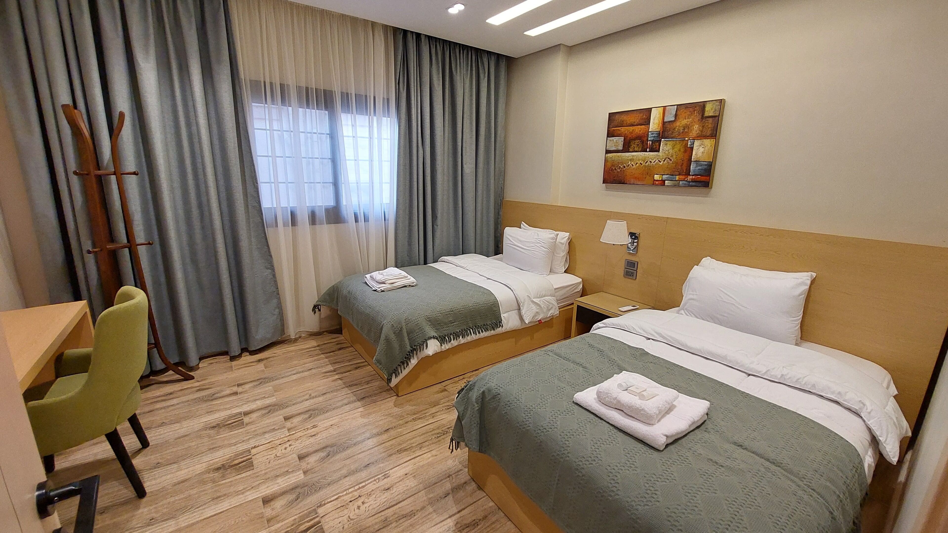 A hotel room with two beds, a desk, a chair, a window with curtains, and a painting on the wall.