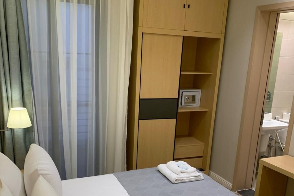 Ένα δωμάτιο ξενοδοχείου με ένα κρεβάτι, ένα κομοδίνο με ένα φωτιστικό, μια ντουλάπα και ένα μπάνιο που φαί νεται μέσα από μια ανοιχτή πόρτα.