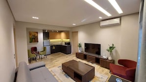 A modern living room with a sofa, coffee table, and TV. There is a kitchen area with cabinets and a refrigerator. - Mirage Taiaa Hotel Apartments (New Cairo)