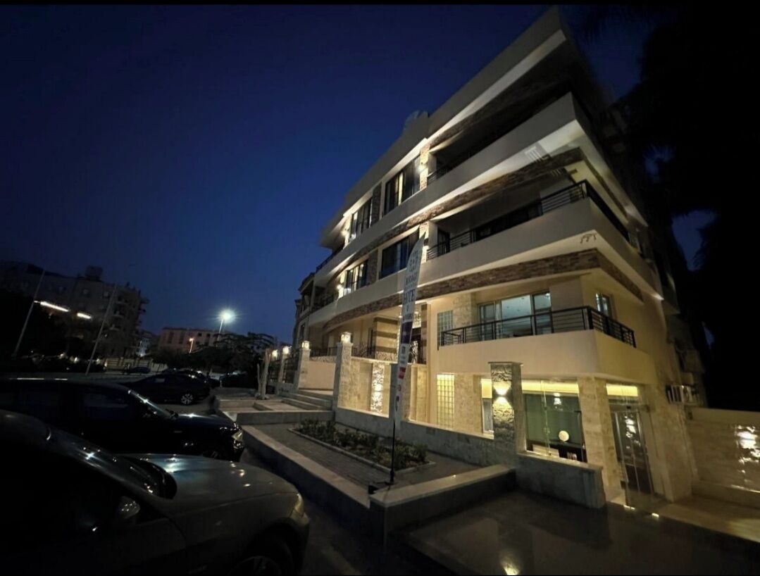 A modern multi-story building with a well-lit entrance and a parking area in front.