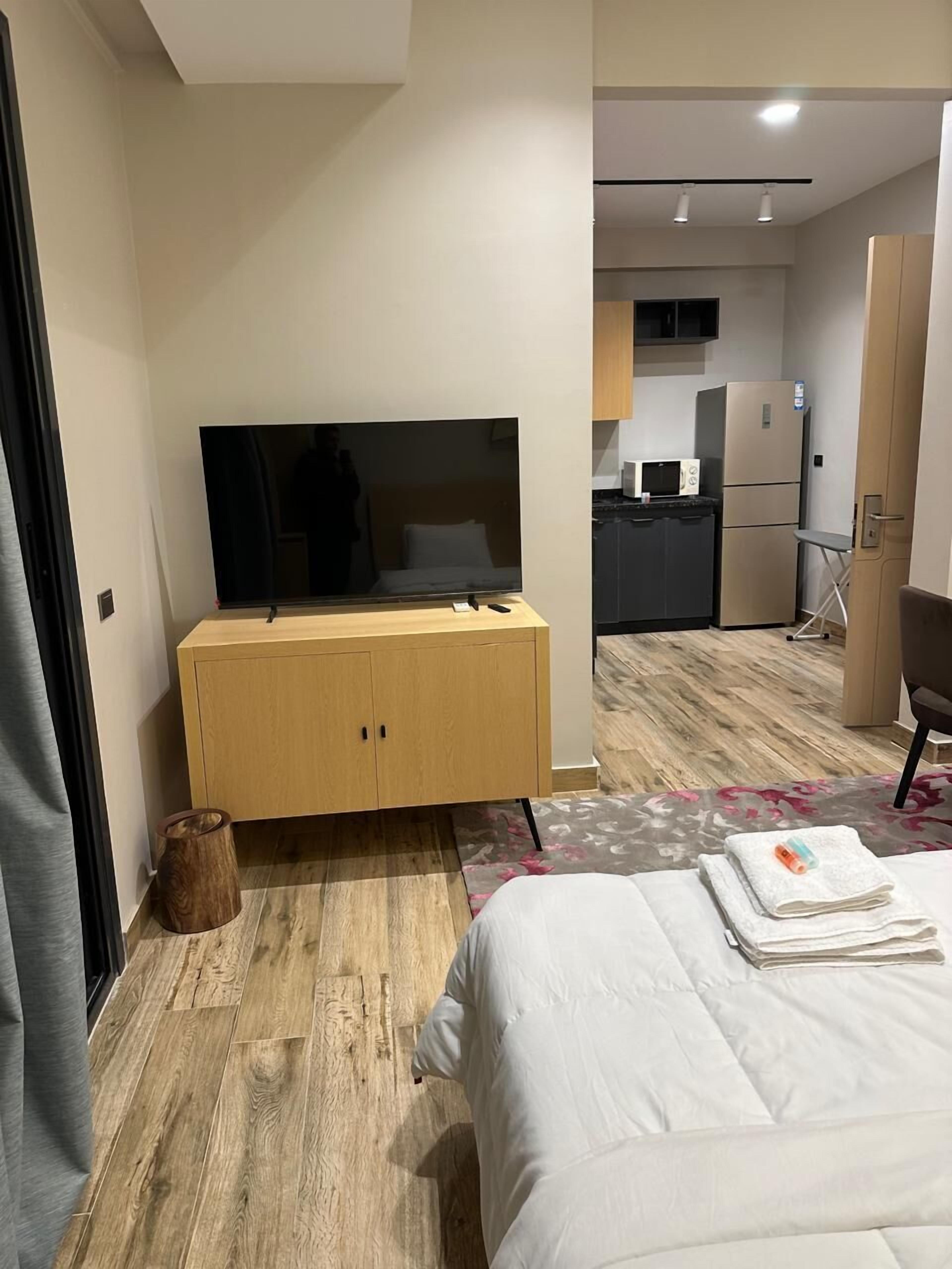 A hotel room with a bed, a wooden dresser, a flat-screen TV, a small kitchen area, and a bathroom.