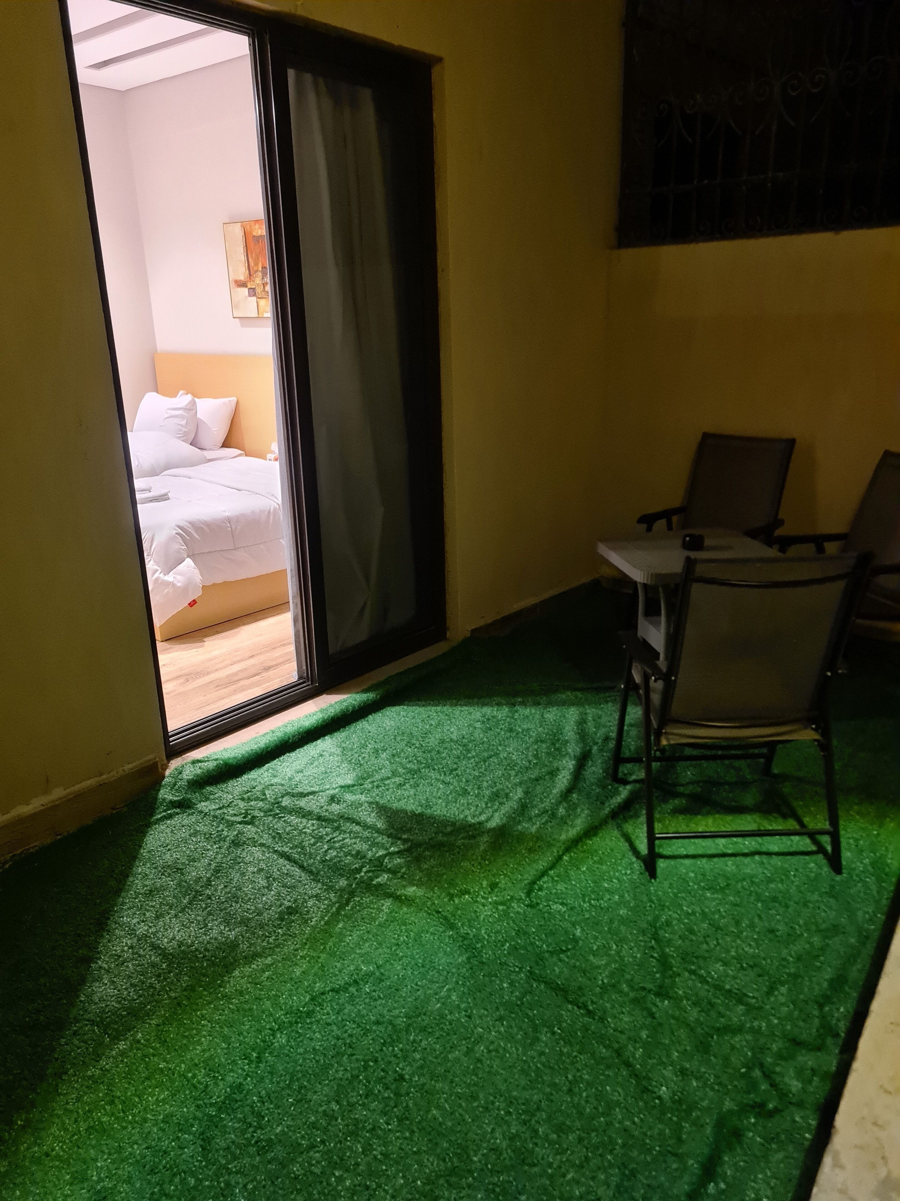 A room with a bed, a green artificial grass floor, a table with chairs, and a sliding glass door leading to another room.