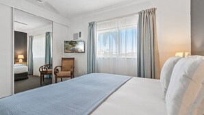Habitación de hotel con cama, una silla, escritorio con lámpara, ventana con cortinas y un cuadro en la pared.