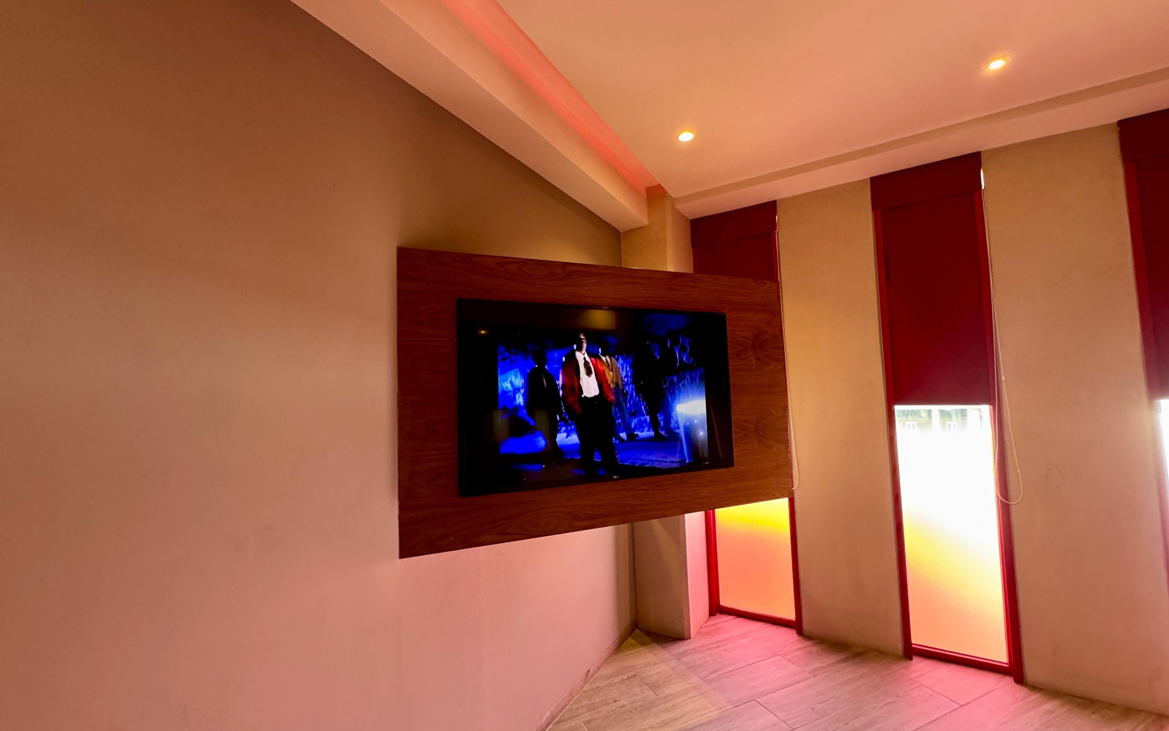 A modern hotel room with a mounted flat-screen TV on the wall, a wooden floor, and a red accent wall.