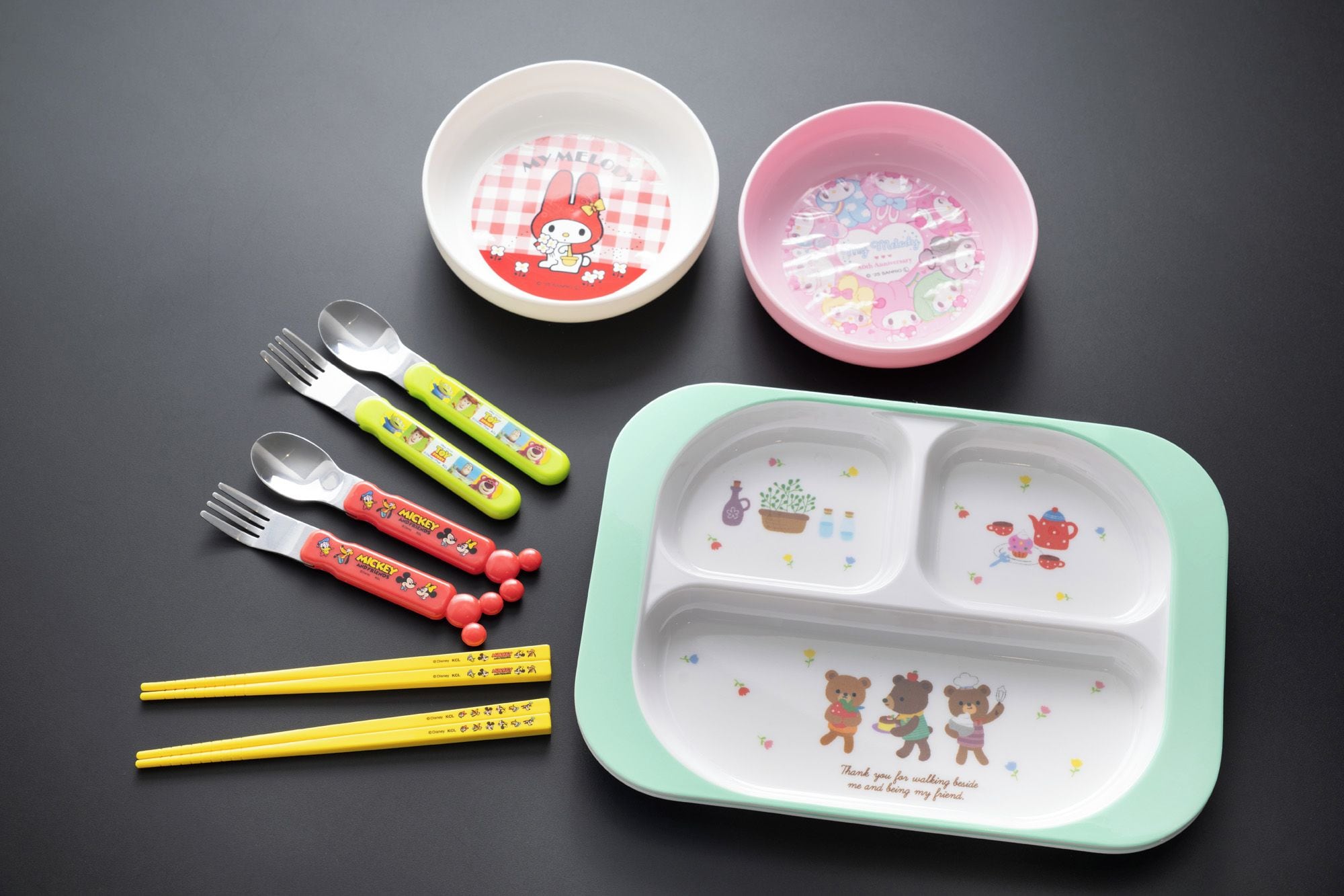 A meal set with a green and white plate, two pink bowls, and several forks and spoons with Mickey Mouse designs.