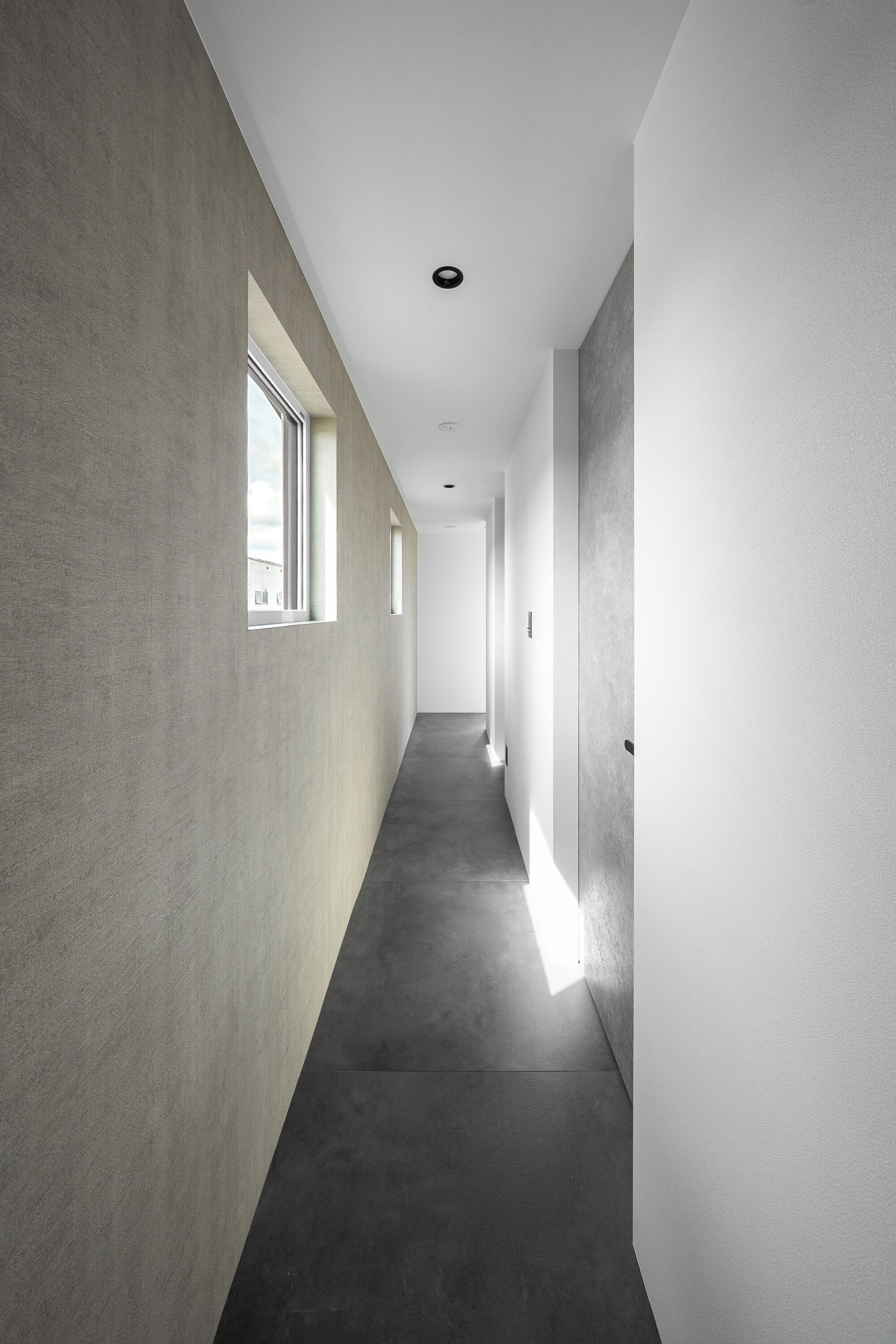 Un pasillo largo y estrecho, con una pared de concreto a un lado y una pared blanca al otro, que conduce a una puerta al final.