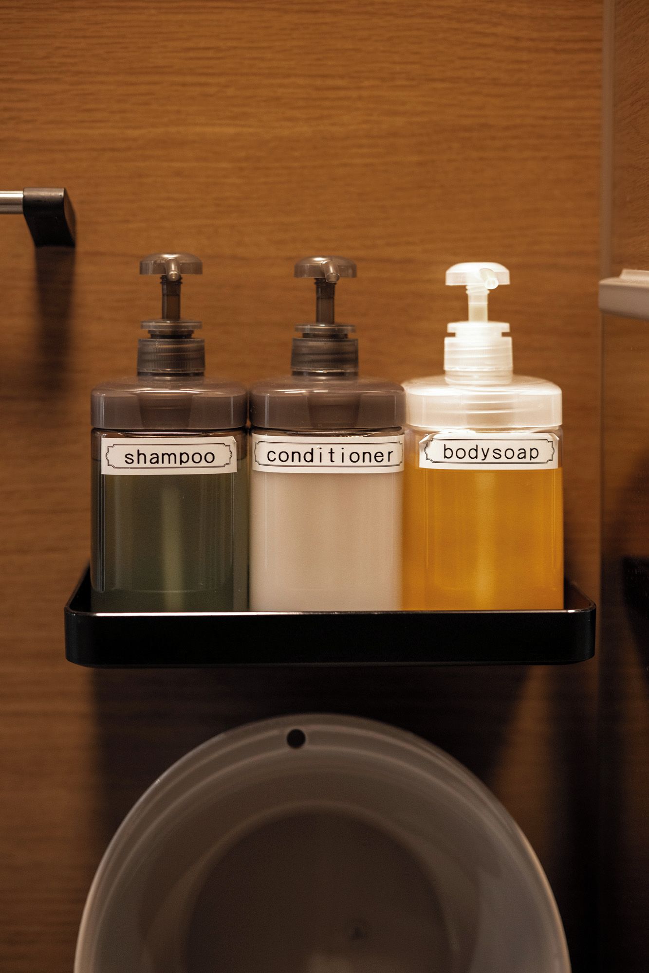 Un estante de baño con tres botellas etiquetadas como champú, acondicionador y jabón de cuerpo.