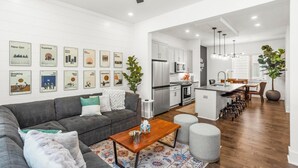 A modern living room with a gray sofa, a wooden coffee table, and a patterned rug. A kitchen with white cabinets and stainless steel appliances is visible in the background.