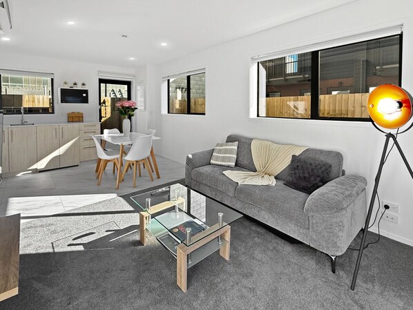 Un salon moderne avec un canapé gris, une table basse en verre et un coin repas avec des chaises blanches.