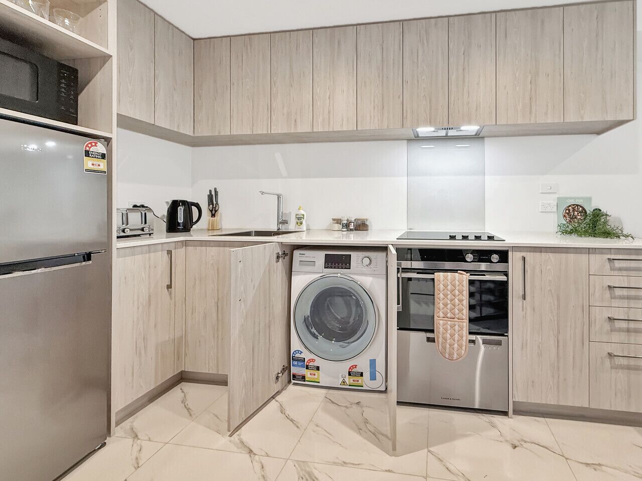 Cocina compacta con lavadora y secadora integradas, electrodomésticos de acero inoxidable y muebles de madera clara.