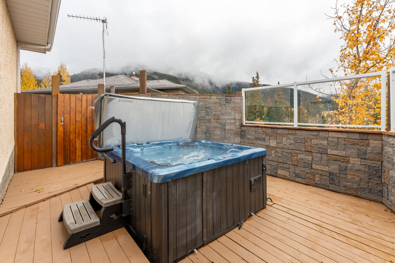 Ein Whirlpool auf einer Holzterrasse mit einer Steinmauer und einem Fenster mit Blick auf ein bewaldetes Gebiet.