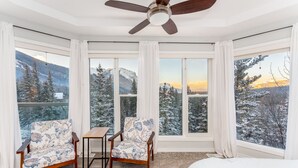 A room with a bed, two chairs, a small table, and a ceiling fan. Large windows offer views of snow-covered trees and mountains. - 4,000 SF Luxury Home | 6-BR + 5-BA | 2-minute DRIVE to Jasper National Park (Jasper)