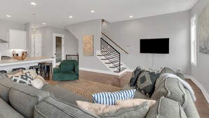 A modern living room with a grey sectional sofa, a green armchair, and a patterned rug. A staircase with a black railing leads to an upper level. - Vibrant 12 Bed Oasis! Luxury meets spacious! Full Kitchen with Huge Rooftop! (Nashville)
