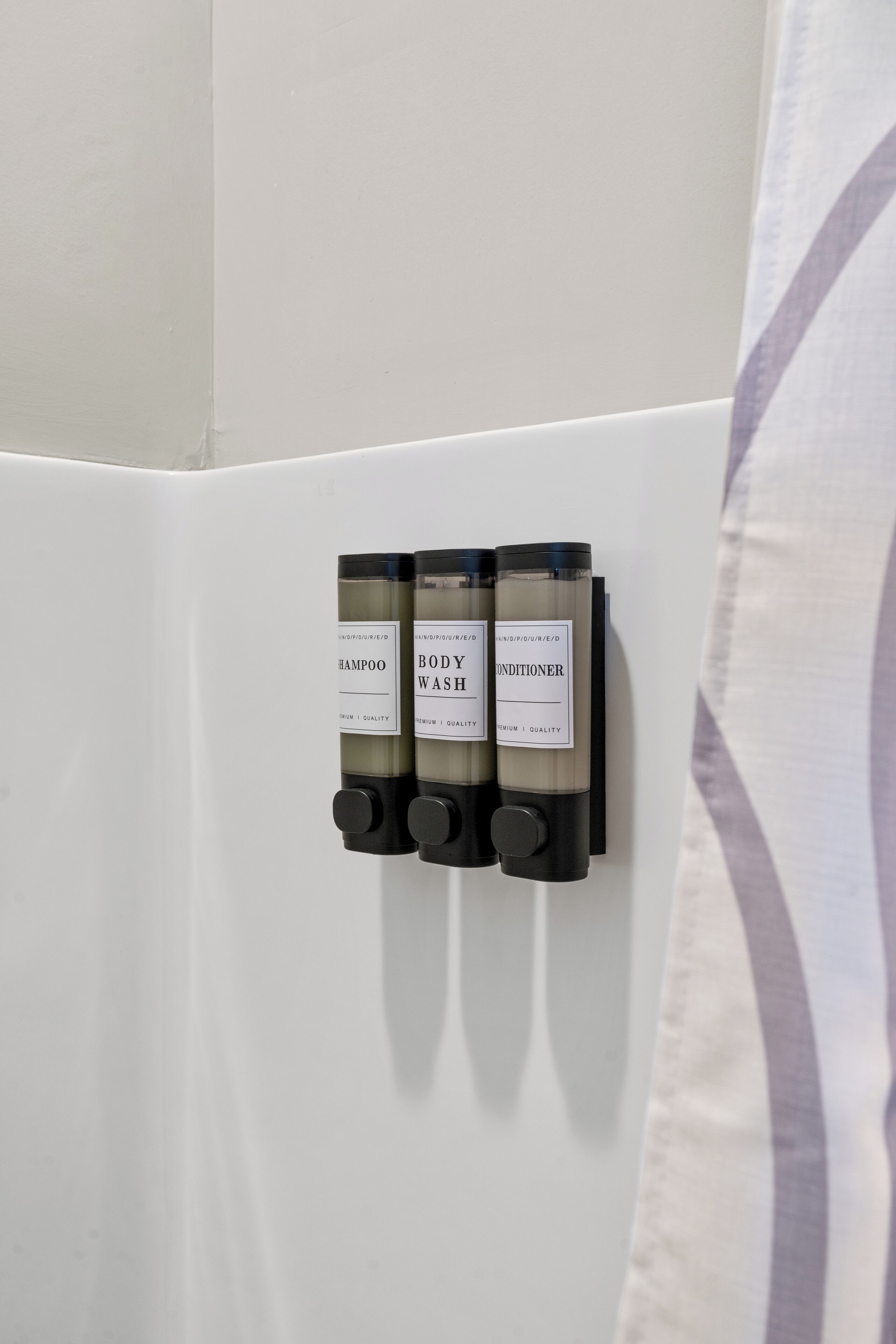 A wall-mounted dispenser with three bottles labeled 'SHAMPOO,' 'BODY WASH,' and 'CONDITIONER.'