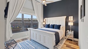 Un dormitorio con un ventanal grande, ventilador de techo, cama con cabecera blanca y ropa de cama azul, mesita de noche con lámpara, y alfombra a rayas azules y blancas.