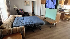 Ein gemütlicher Wohnraum mit Sofa, einem kleinen Holztisch, Deckenventilator, Fernseher und einer Küchenzeile im Hintergrund.