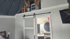 Quarto com teto inclinado, cama com colcha estampada, porta branca que dá para uma área de cozinha e uma planta pequena no teto.