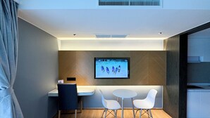 Ein modernes Hotelzimmer mit Holzboden, einem Schreibtisch mit Stuhl, zwei weißen Stühlen, einem an der Wand befestigten Flachbildfernseher und einem Fenster mit Vorhängen.