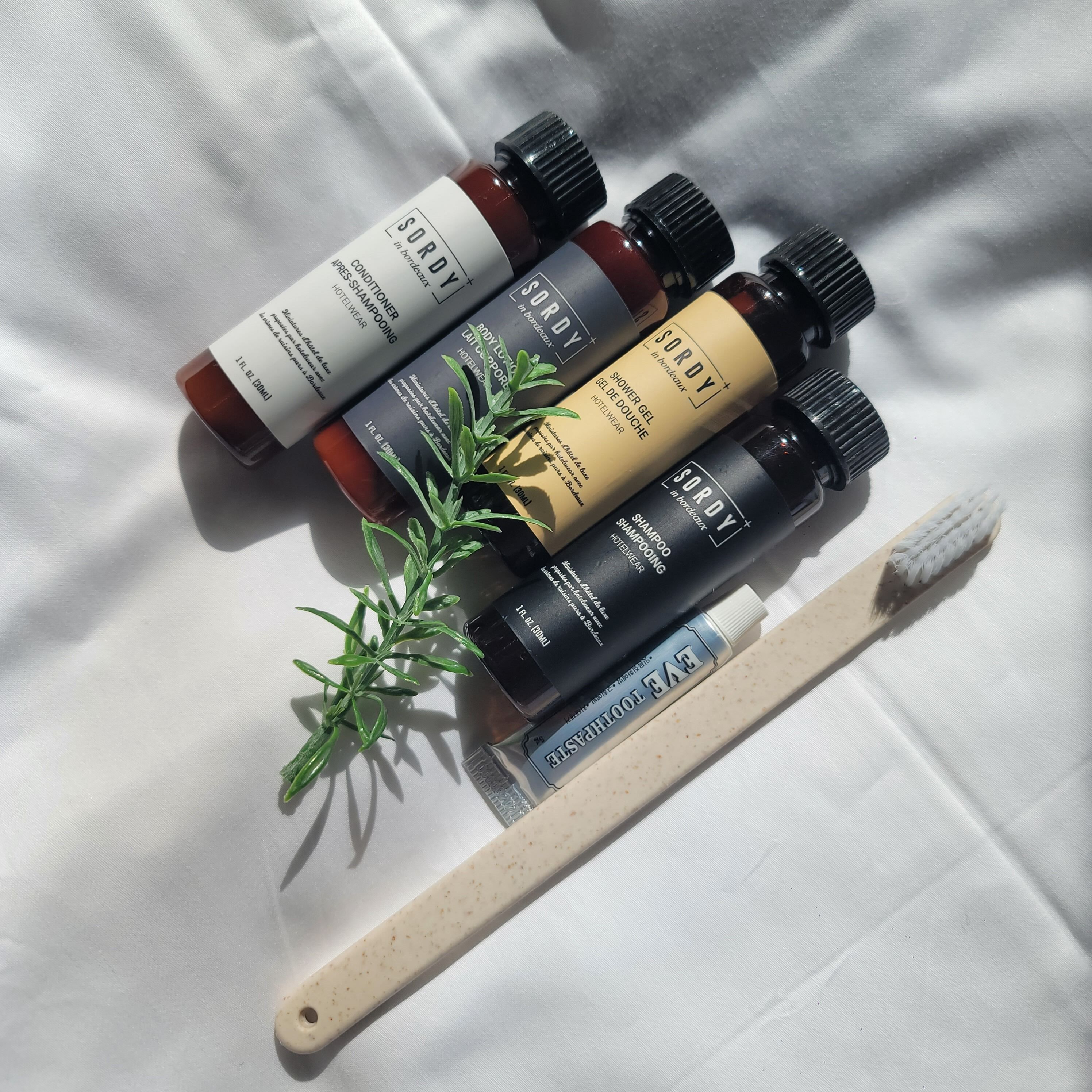 Cuatro botellas de productos de cuidado corporal Sordy, un cepillo de madera y una ramita de romero sobre una superficie de tela blanca.