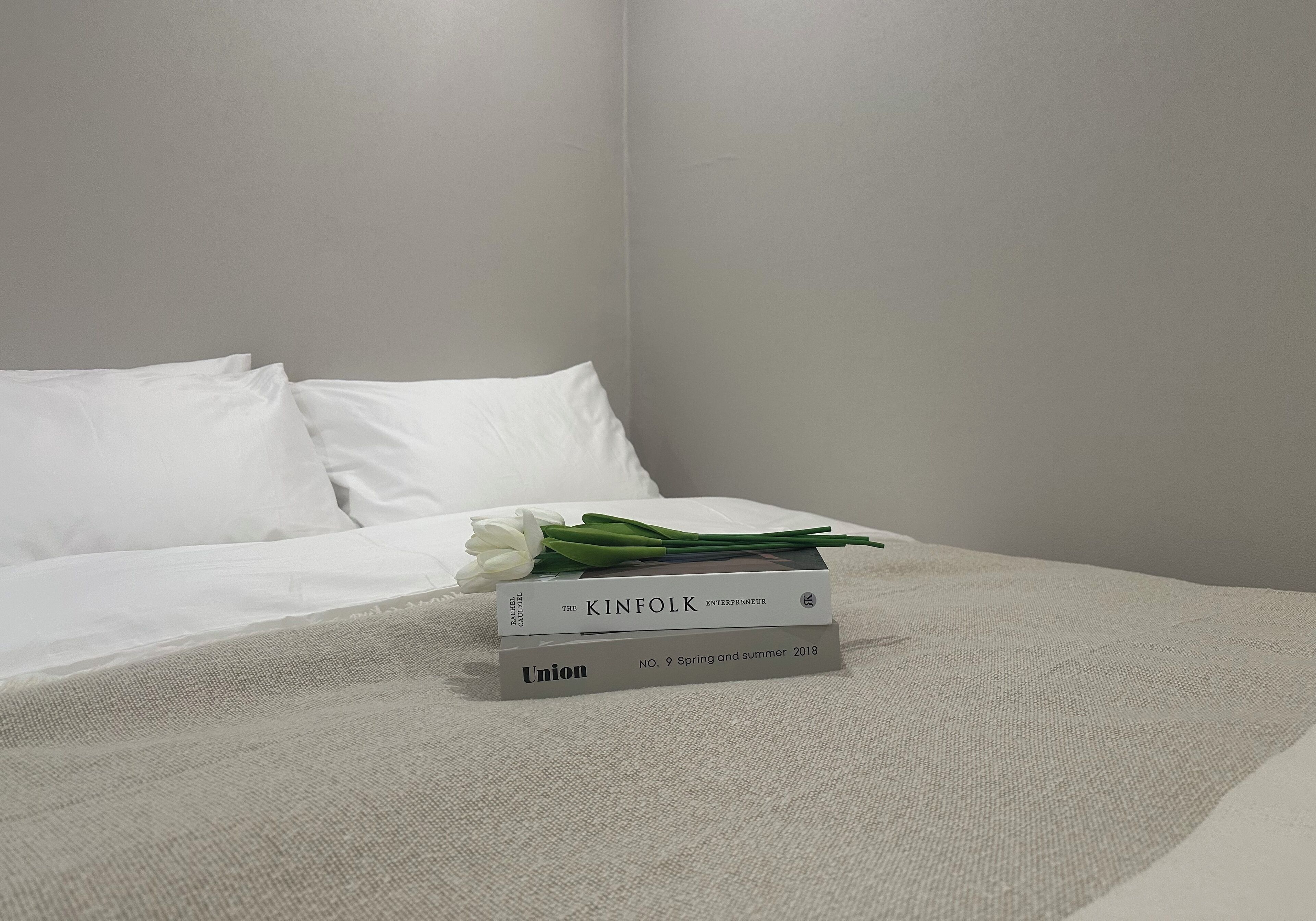 A neatly made bed with white linens and a single white rose on top. There are two books stacked on the bed, one titled "The Kinfolk Entrepreneur" and the other "Union No. 9 Spring and Summer 2018".