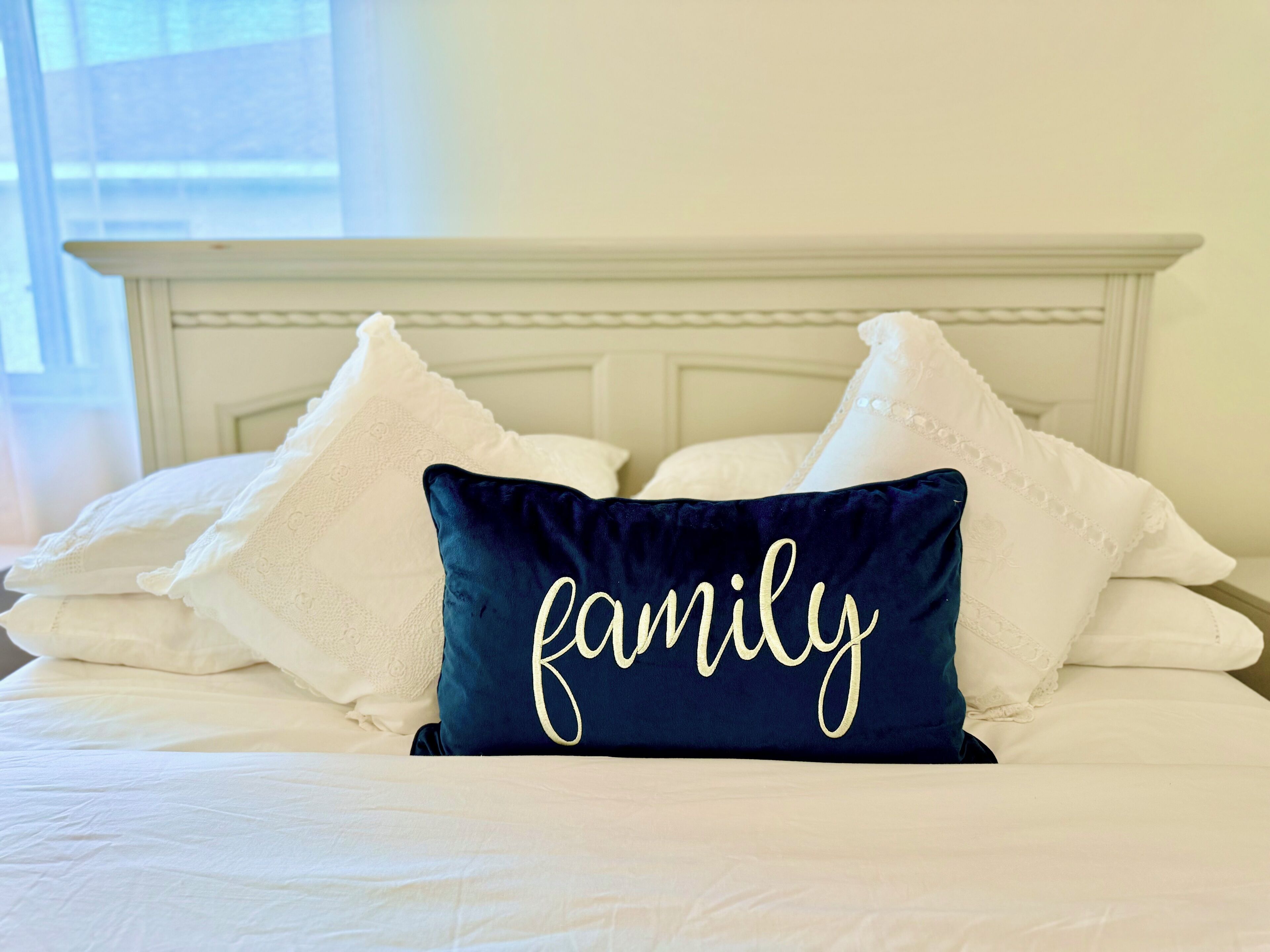 Een bed met witte beddengoed en meerdere kussens, waaronder een marineblauwe kussen met het woord 'family' in gouden letters.