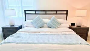 Un lit bien fait, recouvert d’une courtepointe matelassée, avec deux oreillers assortis et une tête de lit en bois. On y trouve deux tables de chevet, chacune avec une lampe, et un smartphone posé sur l’une d’elles.