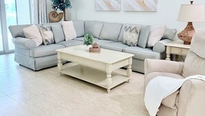 Un salon avec un canapé gris, une table basse blanche et un fauteuil beige. Deux tableaux abstraits ornent le mur, ainsi qu’une plante en pot et une lampe posée sur une table d’appoint.