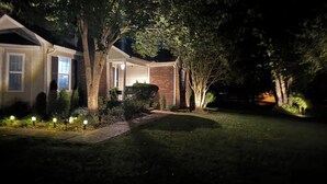 A house with a brick exterior, a covered porch, and a well-lit garden at night. - Convenient, cozy home surrounded by trees, 6 miles to BNA, 13 miles to downtown. (Hermitage)