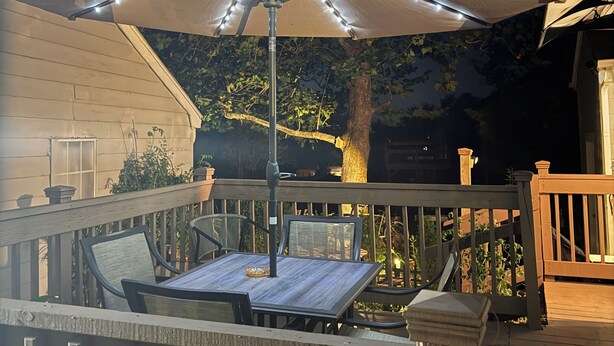 An outdoor patio with a table and chairs, an umbrella with string lights, and a wooden deck. - Convenient, cozy home surrounded by trees, 6 miles to BNA, 13 miles to downtown. (Hermitage)