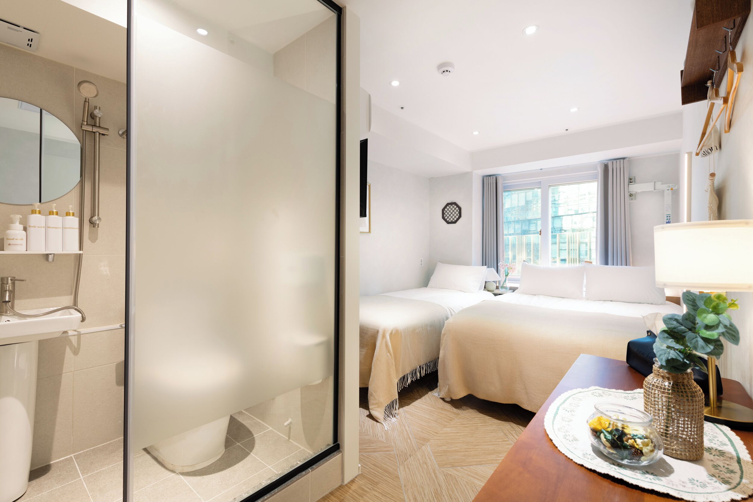 Habitación de hotel con cabina de ducha de vidrio, cama con edredón blanco, mesita de noche con un florero y vistas a la ciudad desde la ventana.