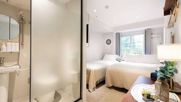 Habitación de hotel con cabina de ducha de vidrio, cama con edredón blanco, mesita de noche con un florero y vistas a la ciudad desde la ventana.