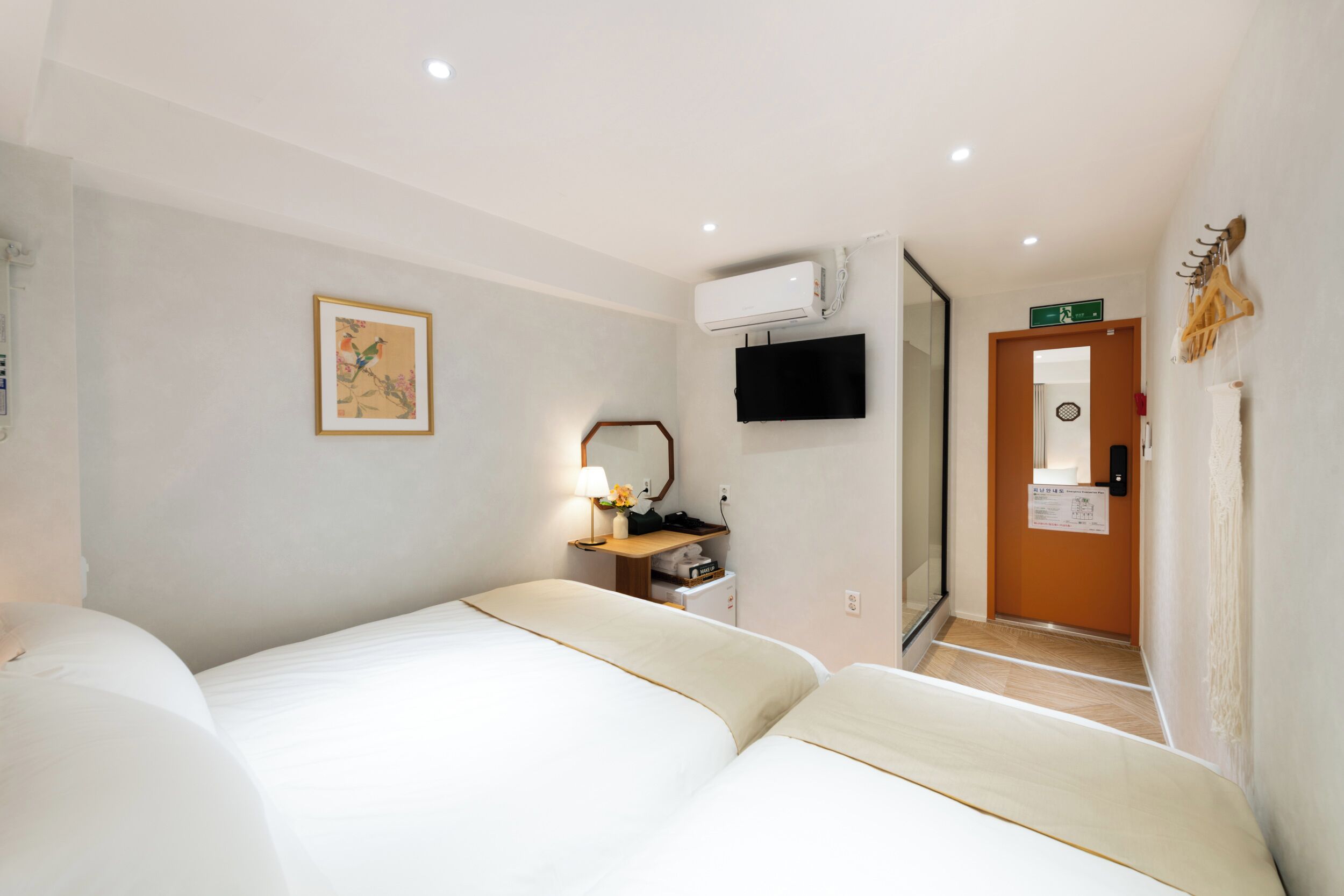 A hotel room with two beds, a flat-screen TV, a wall-mounted air conditioner, a small table with a lamp, and a door leading to another room.