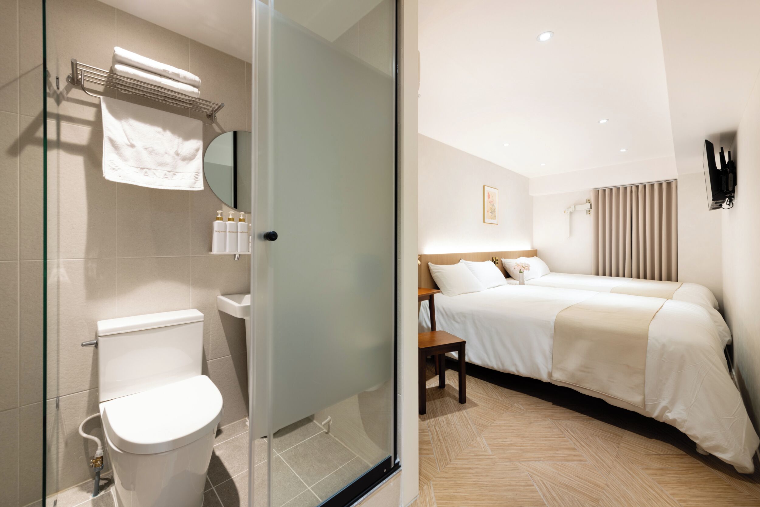 A modern hotel room with a glass shower enclosure, a bed with white bedding, a bedside table, and a mounted TV.