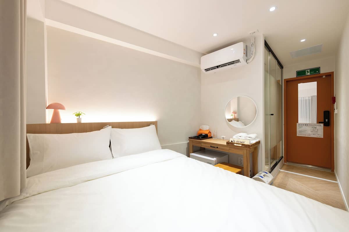 A hotel room with a bed, bedside table, lamp, mirror, and a door with a sign.