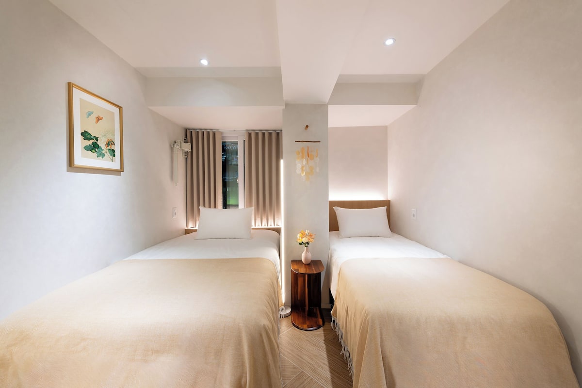 A hotel room with two beds, a framed picture on the wall, a small wooden side table with a vase, and a window with curtains.