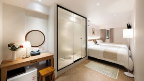 Una habitación de hotel con cama, escritorio con lámpara, espejo, armario y un refrigerador pequeño.