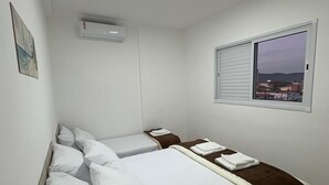 A bedroom with two beds, a window with a view, an air conditioning unit, and a painting on the wall.