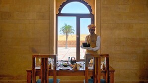 A room with a large arched window, a table set for dining, and a waiter holding a tray. - Desert Valley Resort (Jaisalmer)