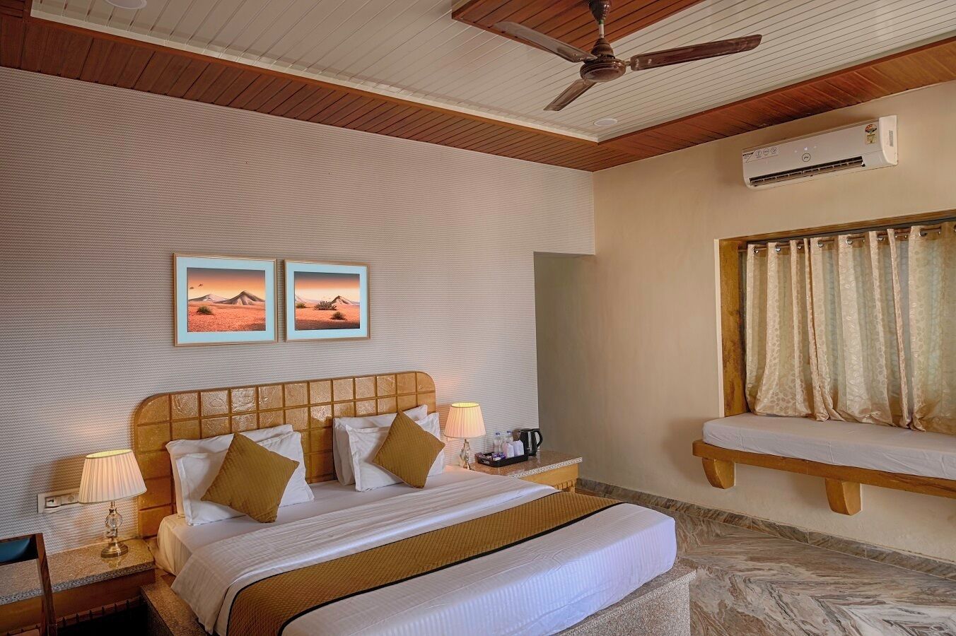 A hotel room with a large bed, two bedside tables with lamps, a ceiling fan, an air conditioning unit, and two framed pictures on the wall.