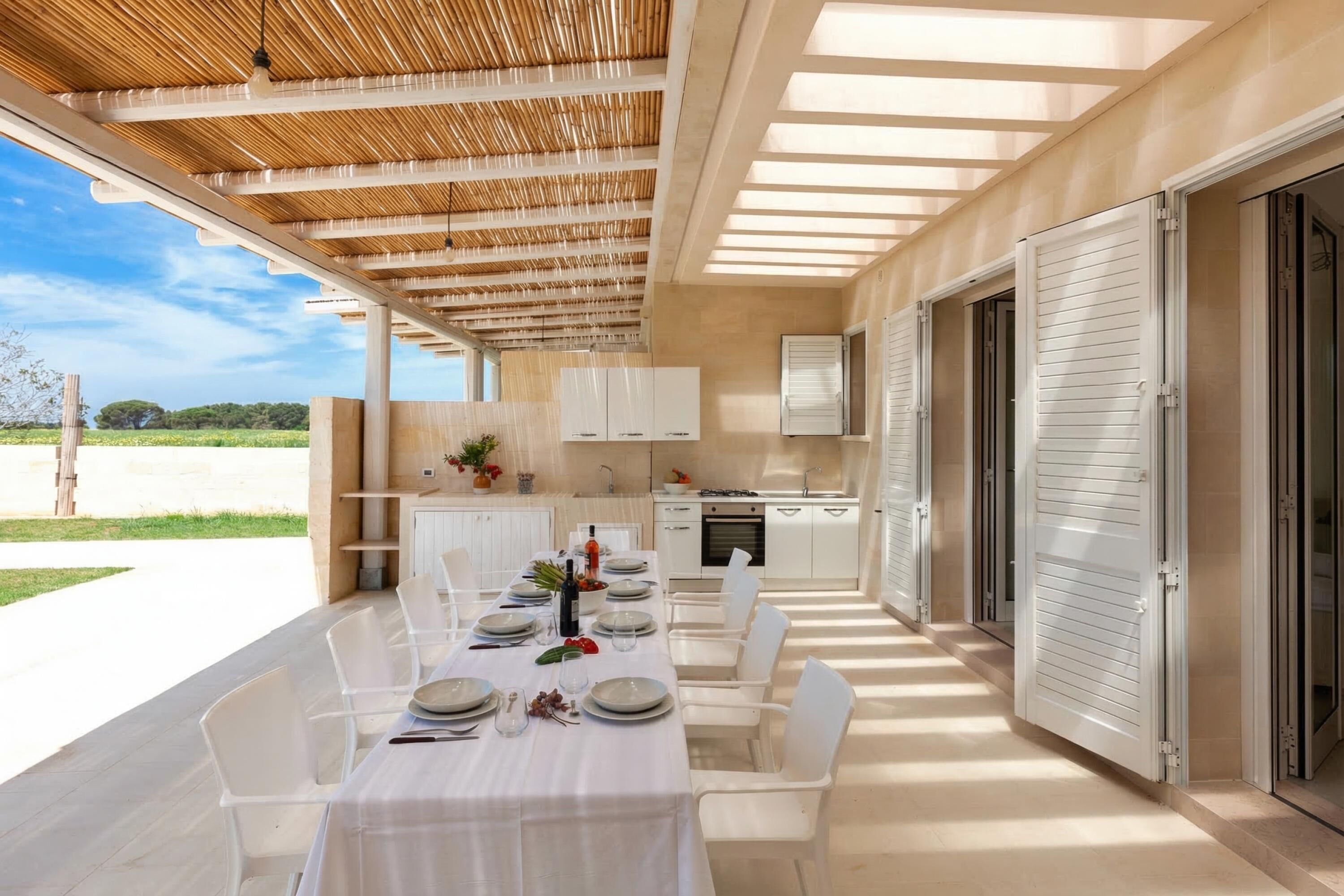 An outdoor dining area with a table set for six, white chairs, a pergola overhead, a kitchen with white cabinets, and a view of the countryside.