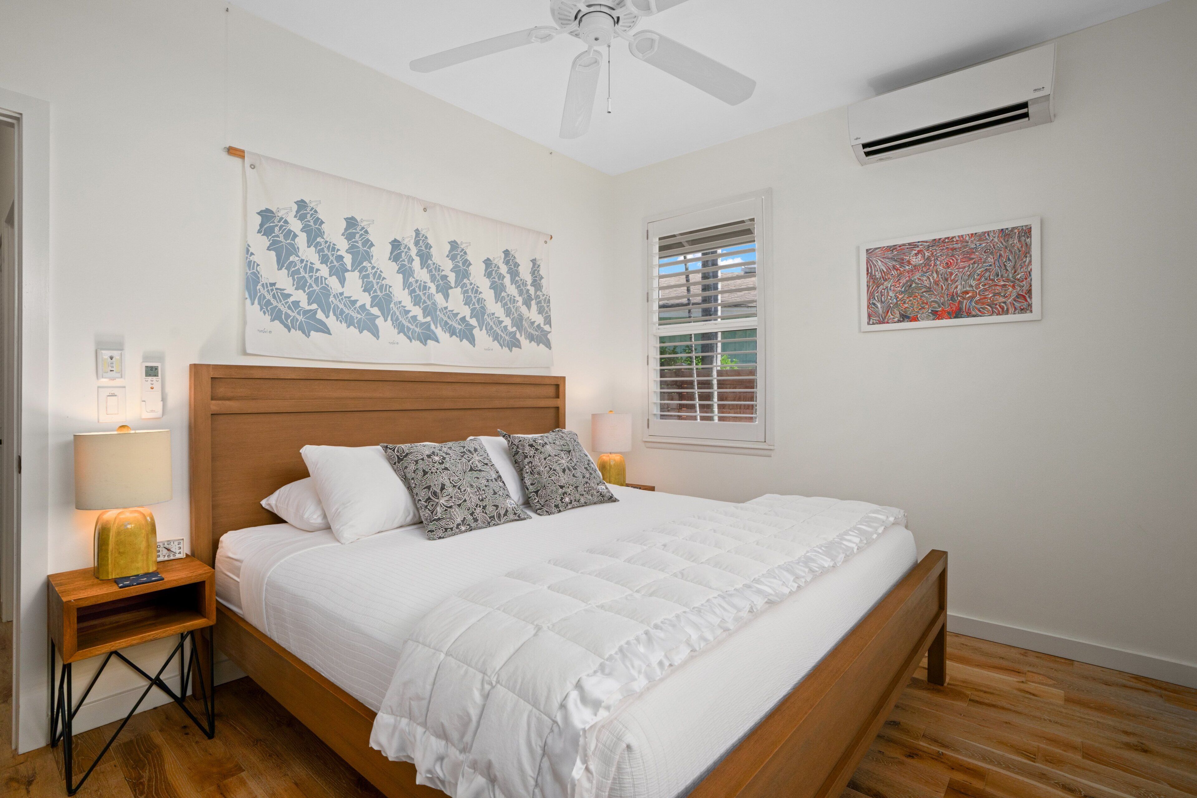 Une chambre avec un lit en bois, du linge de lit blanc, un ventilateur de plafond, un climatiseur, une fenêtre avec des stores et deux lampes fixées au mur.