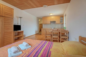 A bedroom with a bed, a TV, wooden furniture, and a kitchenette in the background.