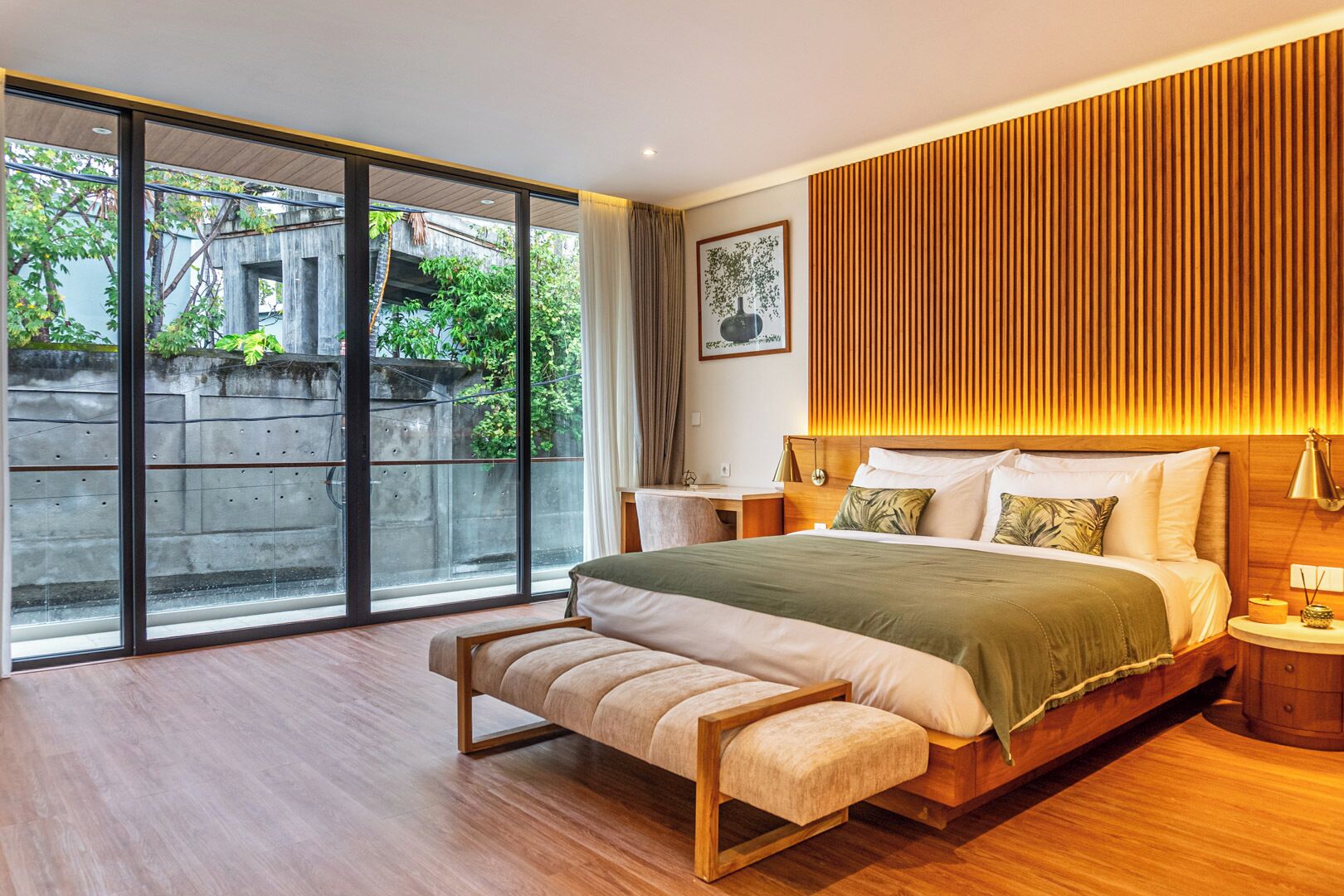 A modern bedroom with a large bed, a bench, a desk, and a view of greenery through large windows.
